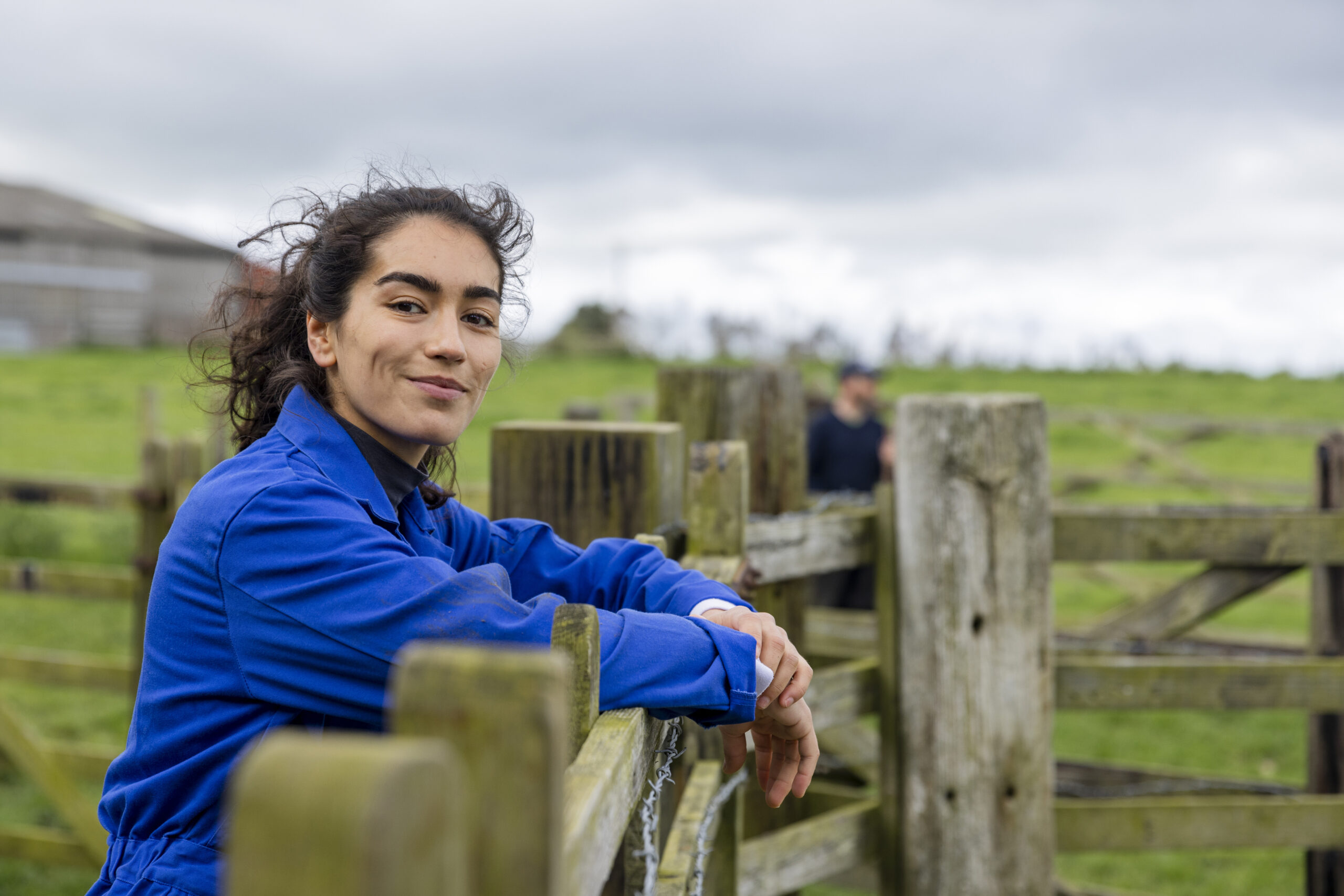 A portrait of a young female farmer wearing overalls, standing and leaning against a fence at the farm she works at in Embleton, North East England while looking at the camera and smiling.